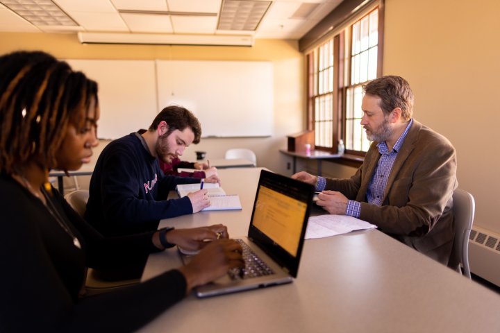A professor sits at a table with two students in a small classroom, engaging in discussion. The students are taking notes, one on a laptop and the other with a notebook, as sunlight streams in through large windows.